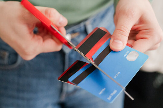 A person in a green sweater is seen cutting a stripe-patterned credit card with red scissors. The act signifies the intention to reduce debt or prevent overspending