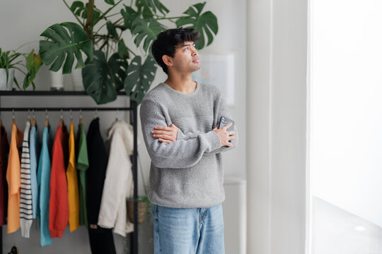 A contemplative young man stands by the window with his arms crossed, looking away thoughtfully. The indoor setting and casual clothing give a relaxed ambiance.