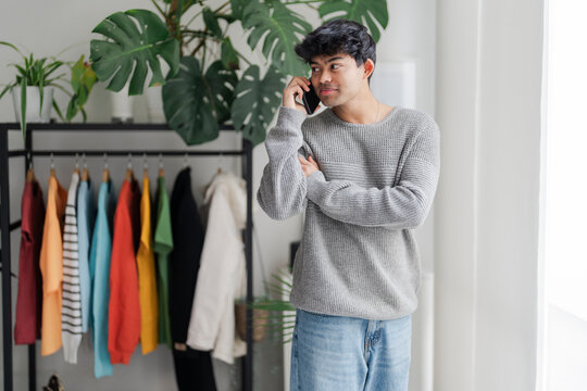 A young man stands by a window, lost in thought while talking on the phone. His casual attire and contemplative demeanor suggest a personal conversation