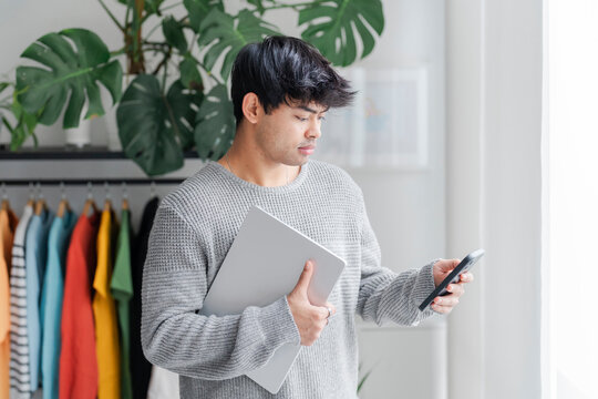 A young man multitasks, holding a laptop in one hand and a smartphone in the other, concentrating on both devices in an indoor setting.