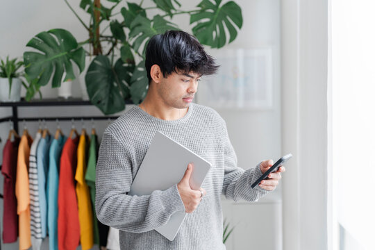 A young man multitasks, holding a laptop in one hand and a smartphone in the other, concentrating on both devices in an indoor setting.