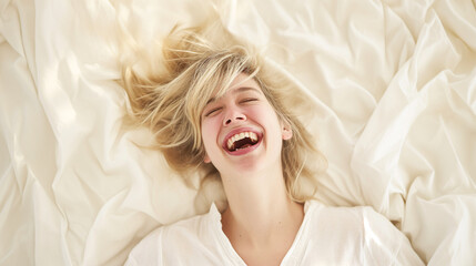 A joyful blonde woman lying on a bed of white linens, laughing and looking up, conveying a carefree and relaxed mood