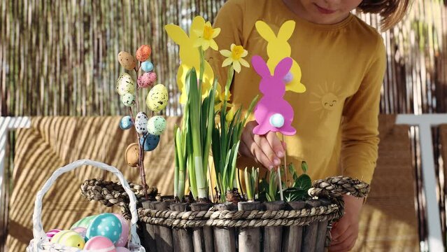 Happy child girl make spring Easter composition with yellow flowers, paper colored Easter bunny, eggs basket. Children prepare decoration present for Easter party at home. Kid creativity and hobby DIY