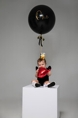 Vertical image of a cute little baby with golden crown sit on little cube and playing with a heart balloon, near flying big black balloon.