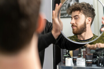 Fototapeta na wymiar Young man adjusting his hairstyle in the mirror - Focused on personal grooming and style.