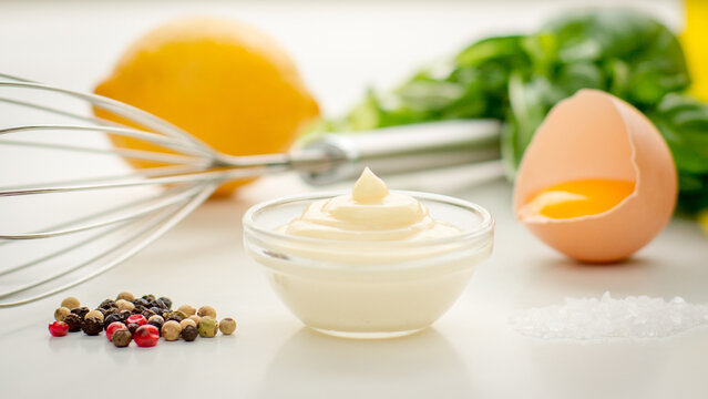 Freshly made mayonnaise with ingredients and a whisk on a white surface