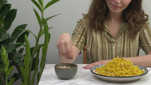 Woman sprinkles the kitchari dish (yellow split mung beans with basmati rice and spices) with chopped cashew nuts. Ayurvedic cleanse food. Potted plant on the side.