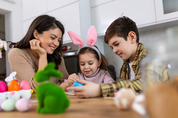 Cute little children wearing funny bunny ears headbands embracing and kissing young happy mother while painting Easter eggs together, sitting at table in kitchen, selective focus