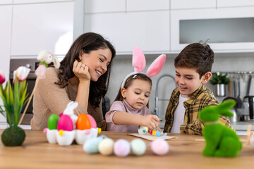 Mother, son and daughters are painting eggs. Happy family are preparing for Easter. Cute little child girl wearing bunny ears. - obrazy, fototapety, plakaty
