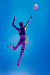 Rear view of young woman, beach volleyball player hits ball in motion against gradient blue background in pink neon light. Movement. Concept of sport, active and healthy lifestyle, power and strength.