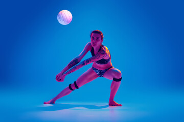 Fototapeta na wymiar Young woman, volleyball player in motion, training, playing against gradient blue background in pink neon light. Concept of sport, movement, active and healthy lifestyle, power and strength.