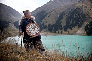A young boy in the viking cosplay costume holding a wooden shield and a axe and standing next to the beautiful alpine lake with Mountains view