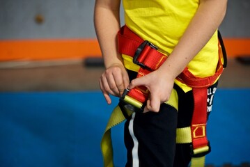 Young European boy in a yellow shirt is putting on his harness next to the climbing wall in an...