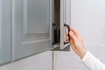 Selective focus on woman hand opening grey wooden door on kitchen cabinet.