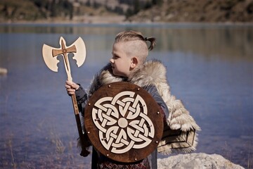 A young boy in the viking cosplay costume holding a wooden shield and a axe and standing next to the beautiful alpine lake with Mountains view