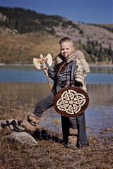 A young boy in the viking cosplay costume holding a wooden shield and a axe and standing next to the beautiful alpine lake with Mountains view