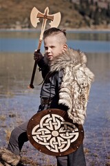 A young boy in the viking cosplay costume holding a wooden shield and a axe and standing next to the beautiful alpine lake with Mountains view
