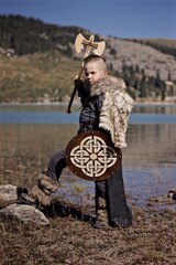 A young boy in the viking cosplay costume holding a wooden shield and a axe and standing next to the beautiful alpine lake with Mountains view