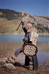 A young boy in the viking cosplay costume holding a wooden shield and a axe and standing next to the beautiful alpine lake with Mountains view