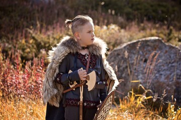A young boy in the viking cosplay costume holding a wooden shield and a axe and standing in the Mountains