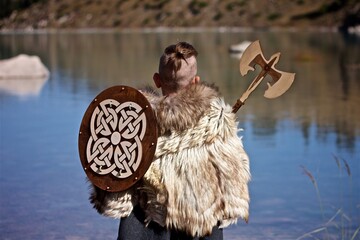Rear view of a young boy in the viking cosplay costume holding a wooden shield and a axe and...