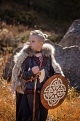 A young boy in the viking cosplay costume holding a wooden shield and a axe and standing in the Mountains 