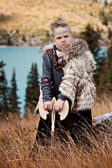 A young boy in the viking cosplay costume holding a wooden axe and standing next to the beautiful alpine lake with Mountains view