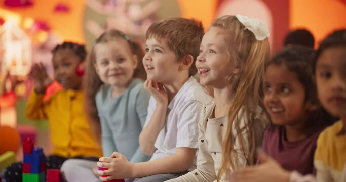 Group of Happy Excited Kids Sitting on a Floor and Watching Cartoons on a TV in a Modern Daycare Center. Diverse Children Having Fun, Learning Through Video Lessons, Games, Music, Art 