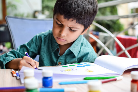 Smart Indian little boy perform thumb painting with different colourful water colour kit during the summer vacations, Cute Indian Kid doing colourful thumb painting drawing on wooden table