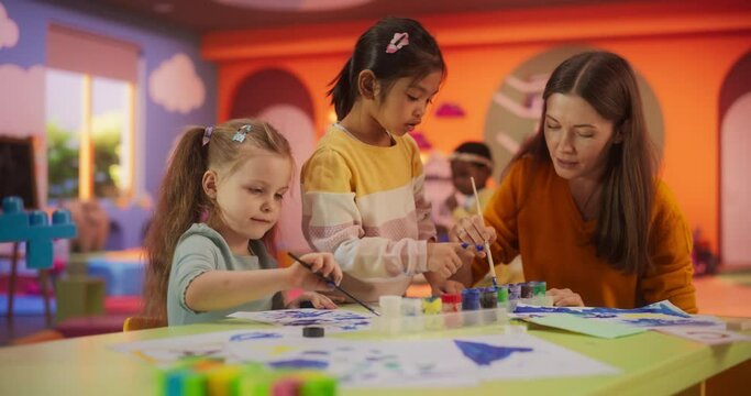 Two Adorable Multiethnic Girls Using Watercolor to Create Colorful Paintings on Paper. Cheerful Female Teacher Spending Productive Time in Preschool, Teaching Kids to Paint in Art Class
