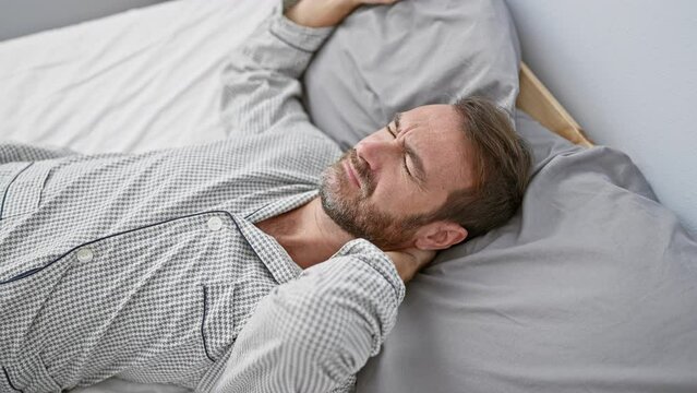 Middle-aged hispanic man in pajamas feeling neck pain while lying in bed indoors at home, portraying discomfort and health issues.