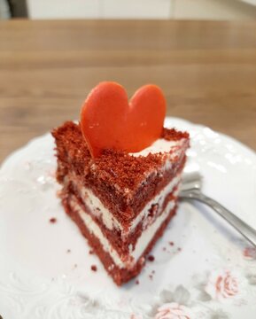 A piece of cake on a dessert plate made from layers of red sponge cake with cream and a heart on top. Red velvet cake with red chocolate heart.