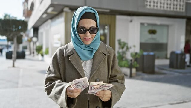 Mature woman with headscarf counting saudi riyal banknotes on urban street.