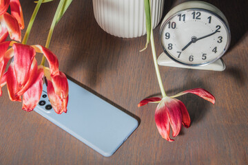 Still life of the theme of loneliness. Withered flowers in a vase, a table clock and a smartphone left on the table