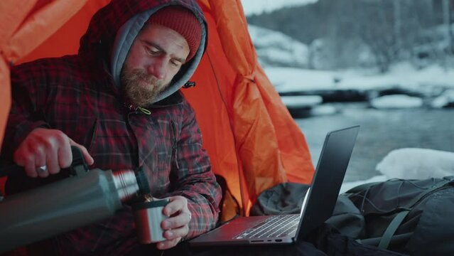 Man sitting in camping tent, pouring hot tea from thermos into cup, drinking and using laptop during winter travel in nature