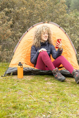 Curly and blonde hair, mountaineer woman camps with his orange tent and using his smart phone