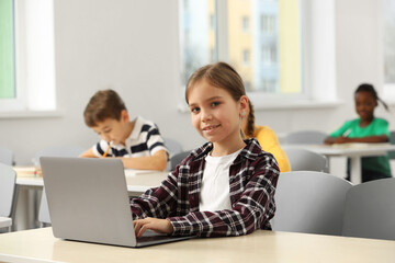 Smiling little girl with laptop studying in classroom at school
