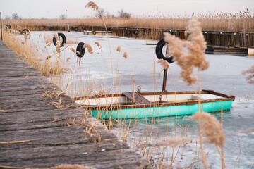 A turquoise wooden rowboat moored in winter on the frozen lake amidst the reeds. A wooden dock...