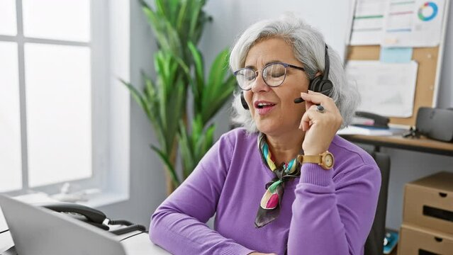 A mature woman with grey hair wearing a headset while working at her office desk.