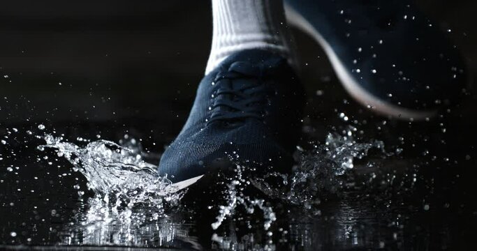 Super slow motion macro of active sportsman wearing sneakers shoes is running through puddle of rainwater with splashing drops jogging workout on street at 1000 fps.