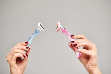 unknown young woman with nail polish holding blue and pink razors in her hands on gray background
