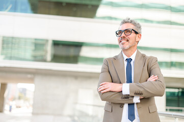 Fototapeta premium Senior businessman stands confidently with arms crossed in front of a modern office building, his expression content and optimistic, embodying success and experience. Leadership and career achievement