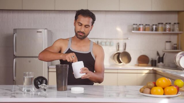 Middle aged Indian man preparing protein shake by adding scoop of powder at kitchen after workout - concept of diet supplement, bodybuilding and fitness routine