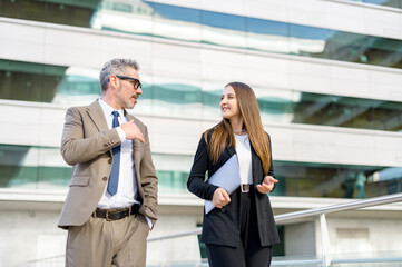 The experienced businessman gestures while talking with his female colleague outdoors, a senior man and young woman in business attire walking and conversing with modern office on background