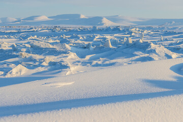 Winter arctic landscape. Snow-covered ice floes on the surface of the frozen estuary. Ice hummocks and snowdrifts. Mountains in the distance. Cold frosty winter weather. Chukotka, Far North of Russia.
