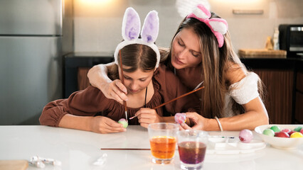 Mom helps her daughter color eggs for Easter. Family in hair holders with bunny ears preparing for...