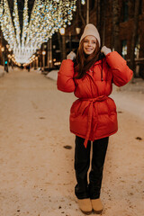 Full length vertical portrait of charming young woman in hat and winter jacket standing posing on snow city street on blurred background of hinged festive light illumination, smiling looking at camera