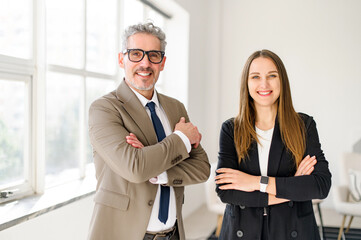 A charismatic senior entrepreneur with grey hair stands arms crossed next to a young professional...