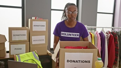 Focused woman volunteer organizes donations indoors at a charity center room with cardboard boxes and clothes.