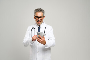 A senior doctor with a friendly demeanor is holding a smartphone, reviewing medical information or scheduling appointments, with a stethoscope draped around his neck and a look of concentration.